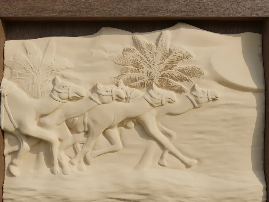 A carved relief panel in cream stone showing a line of camels walking past palm trees, mounted in a dark wooden frame that hangs from two wires against a grey background.