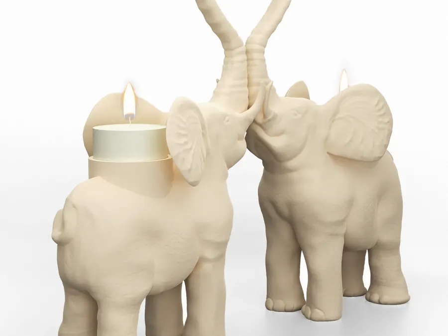 Two cream ceramic elephant figurines facing each other with their trunks raised and curled into a heart shape, each carrying a lit tealight candle on its back, on a plain white surface.