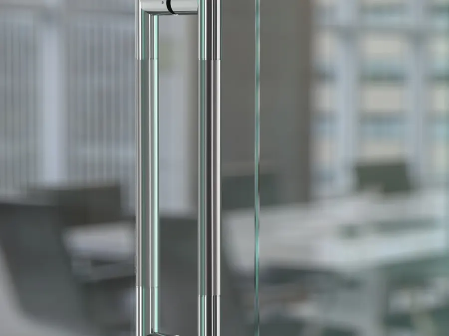 Back to back stainless pull handles on a clear glass door, sharp in the foreground, with a blurred open plan office of desks, chairs and bright windows behind.